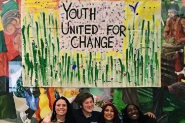 Four Transitional age youth sitting under a painted banner that reads "Youth United for Change"